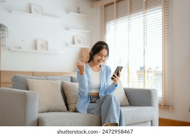 Smiling pretty young Asian woman sitting on cozy couch, using funny mobile apps in living room. Woman at Home, Doing Online Shopping, Messaging Friends, Posting on Social Media - Powered by Shutterstock