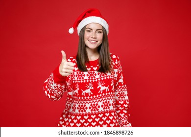 Smiling pretty young brunette Santa woman in sweater Christmas hat showing thumb up looking camera isolated on red colour background, studio portrait. Happy New Year celebration merry holiday concept