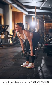 Smiling pretty young female is training legs with dumbbells and doing deadlift in sport club