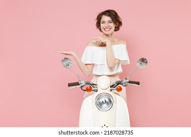 Smiling pretty young woman 20s in white summer clothes posing pointing index finger hand aside on mock up copy space sitting driving moped isolated on pastel pink colour background studio portrait - Shutterstock ID 2187028735