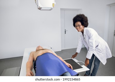 Smiling Professional African American Woman Doctor In Uniform Posing To Camera, While Taking Chest Xray Of Male Caucasian Patient. X-ray Scan Of Lungs And Skeleton In Modern Clinic