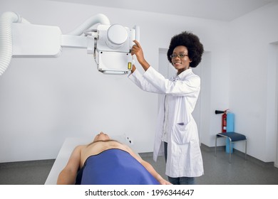 Smiling Professional African American Woman Doctor In Uniform Posing To Camera, While Taking Chest Xray Of Male Caucasian Patient. X-ray Scan Of Lungs And Skeleton In Modern Clinic