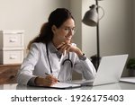 Smiling professional female doctor wearing glasses and uniform taking notes in medical journal, filling documents, patient illness history, looking at laptop screen, student watching webinar