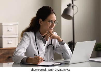 Smiling professional female doctor wearing glasses and uniform taking notes in medical journal, filling documents, patient illness history, looking at laptop screen, student watching webinar