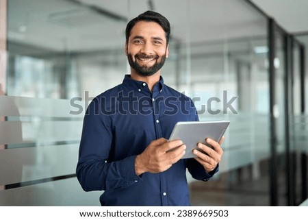 Smiling professional Indian businessman entrepreneur using tab computer working standing in office. Confident business man holding digital tablet tech device working, looking at camera. Portrait.
