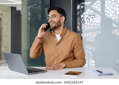 Smiling professional man at his office desk, having a phone call, with a laptop and tablet in front of him. - Powered by Shutterstock