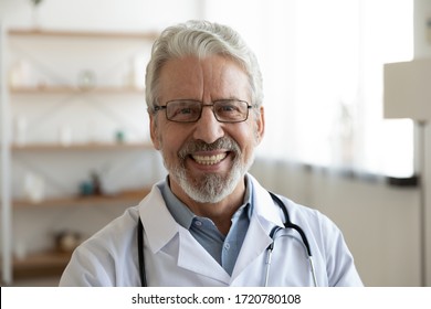 Smiling Professional Older Man Doctor Wears White Coat, Glasses And Stethoscope Looking At Camera. Happy Bearded Senior Physician Or Therapist With Dental Smile Posing For Close Up Head Shot Portrait.