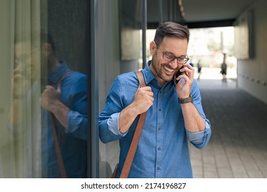 Smiling Professional Talking On Mobile Phone. Young Businessman Is Carrying Laptop Bag. He Is Reflecting On Window Of Office Building At The City.