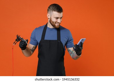 Smiling professional tattooer master artist man in t-shirt apron hold machine black ink in jar, equipment for making tattoo art on body using mobile cell phone isolated on brown background studio