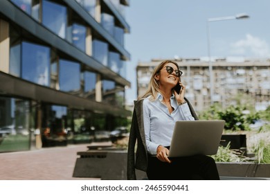 A smiling professional using her smartphone and laptop outside a sleek, contemporary office building on a sunny afternoon.
