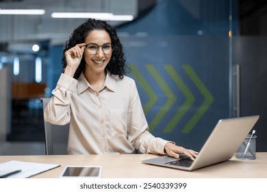 Smiling professional woman adjusting glasses while working at her desk. Laptop and smartphone on table. Ideal image for business, technology, or office-related content. - Powered by Shutterstock