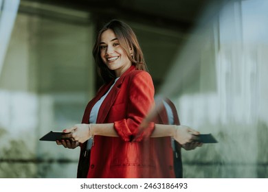 A smiling professional woman stands near a modern glass office building, holding a tablet and looking confident during the evening. - Powered by Shutterstock