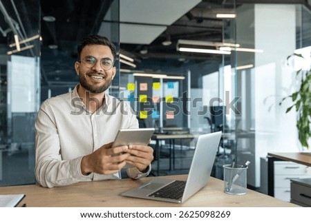 A smiling programmer holding a tablet sits at a desk with a laptop. He looks happy and content in his modern office setting