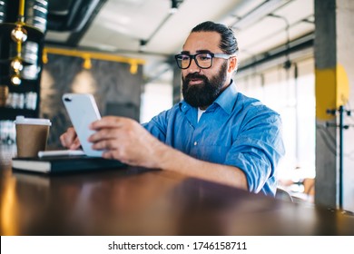 Smiling Prosperous Caucasian Male Owner Of Small Business Sitting Near Bar Checking Mail On Mobile Phone, Positive Maturen 40 Years Man Reading News And Share Media Using Smartphone Connected To 4G