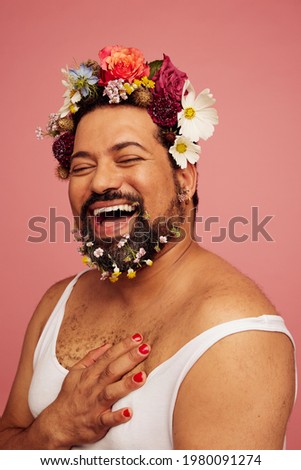 Smiling queer wearing makeup and tank top. Cheerful drag queen wearing flowers in his head and beard.