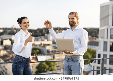 Smiling real estate agent presenting new home keys to happy client outdoors. Woman giving thumbs up gesture and man holding laptop, urban setting with cityscape in background. - Powered by Shutterstock