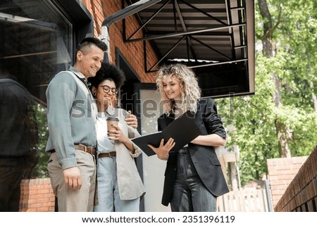 smiling real estate agent showing documents to interracial couple with paper cup near cottage