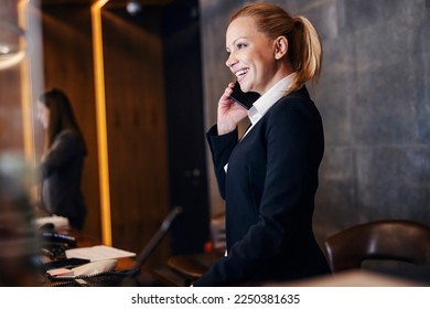 A smiling receptionist is talking on the phone with guest and making a reservation at hotel reception.