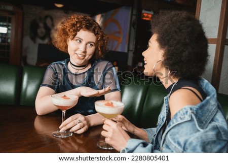 Smiling red haired woman talking to african american girlfriend near cocktails in bar