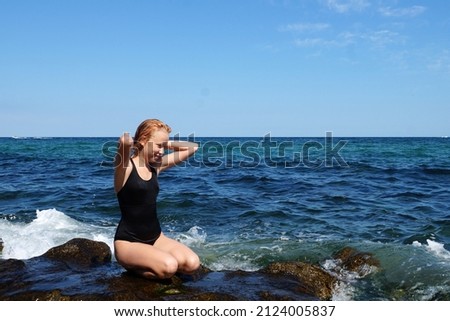 smiling red-haired teen girl in a black swimsuit sits on the seashore against the background of the horizon.