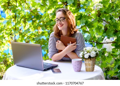 Smiling red-haired woman working on a laptop and holding a notebook. A modern concept of remote work, a freelancer, a digital nomad. - Shutterstock ID 1170704767