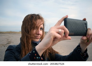 Smiling redhead woman makes photo on her smartphone on a sandy beach in autumn, selective focus, filter applied. Lady in black leather jacket and dotted dress takes pictures on her smartphone on beach