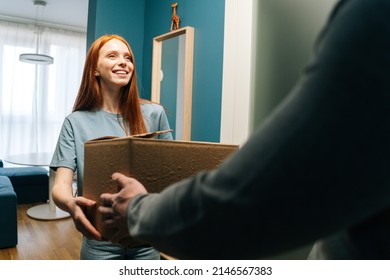 Smiling redhead young woman accepting cardboard box from unrecognizable delivery man on doorstep at apartment. Rear view of courier male delivering parcel package post to female client at home.