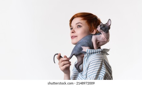 Smiling Redhead Young Woman With Canadian Sphynx Cat Sitting On Her Shoulder. Portrait Of Hipster Dressed In Striped White-blue Shirt. Studio Shot On White Background, Copy Space. Part Of Series.