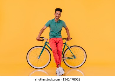 Smiling Refined Guy With Black Hair Posing With Pleasure Near Bike. Indoor Portrait Of Enthusiastic African Man With Green Bicycle.