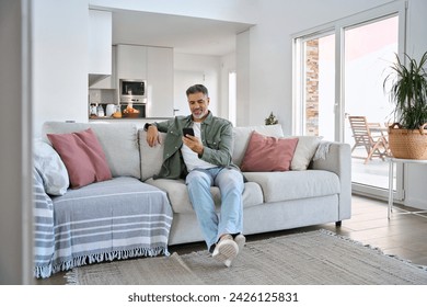 Smiling relaxed senior mature middle aged man holding cell mobile phone using smartphone sitting at home on couch in modern living room, scrolling social media, buying online, texting messages. - Powered by Shutterstock