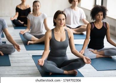 Smiling relaxed young caucasian woman enjoying group yoga training with happy multiracial people indoors, sitting barefoot on mat in lotus padmasana pose, meditating or doing breathing exercises. - Shutterstock ID 1769702306