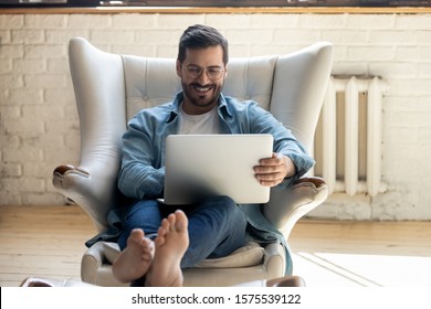 Smiling Relaxed Young Man Sit On Comfortable Armchair Using Laptop Notebook Relaxing Laughing Watching Funny Movie, Happy Guy Enjoy Leisure Time Indoor With Computer Having Fun Online In Social Media