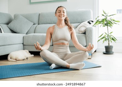 Smiling and relaxed young woman doing yoga exercises and stretches sitting on the floor at home in lotus pose. Concept of healthy lifestyle and home training. - Powered by Shutterstock