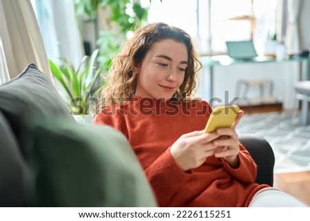 Smiling relaxed young woman sitting on couch using cell phone technology, happy lady holding smartphone, scrolling, looking at cellphone enjoying doing online ecommerce shopping in mobile apps.