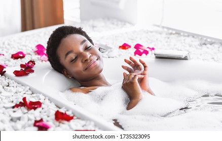 Smiling and relaxing african american woman with closed eyes takes bath with foam and petals