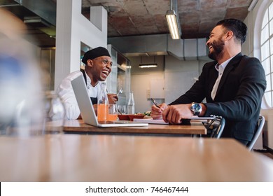 Smiling Restaurant Manager Sitting At Table And Talking With Chef. Restaurant Owner Having A Friendly Conversation With Employee.