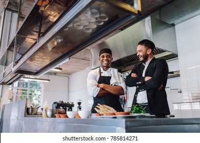 Smiling Restaurant Owner And Chef Standing In Kitchen. Businessman With Professional Cook Standing Together And Laughing.