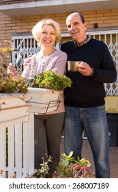 Smiling Retired Couple Outdoors Outside Their Home In A Patio. Husband Hugs Hus Wife And Holds A Cup Of Coffee