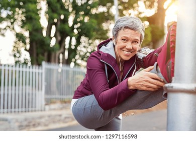 Smiling retired woman listening to music while stretching legs outdoors. Senior woman enjoying daily routine warming up before running. Sporty lady doing leg stretches and looking at camera. - Powered by Shutterstock