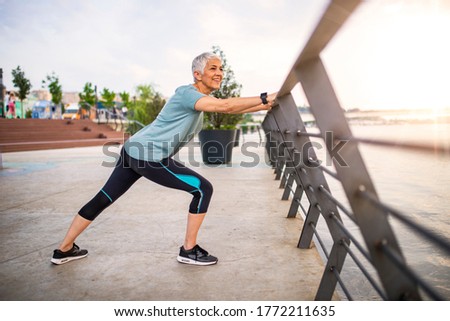 Smiling retired woman stretching legs outdoors. Senior woman enjoying daily routine warming up before running at morning. Sporty lady doing leg stretches before workout 