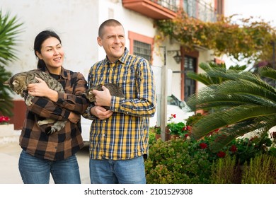 Smiling Romantic American Man And Asian Woman Standing With Their Pets Cats Near Home At Street