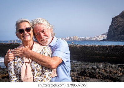 Smiling romantic senior couple with white hairs hugging at the beach looking at camera - horizon over water and seascape - concept of active lifestyle for elderly people in holidays - Powered by Shutterstock