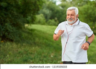 Smiling running old man in green city park. Smiling, feeling good. Sport lifestyle. Wearing classic white polo shirt with dark blue stripes, sport watch, red headphones. Trees, grass on background.