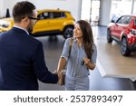 Smiling salesman or manager giving car keys and shakes hand to modern happy woman new car owner in car dealership