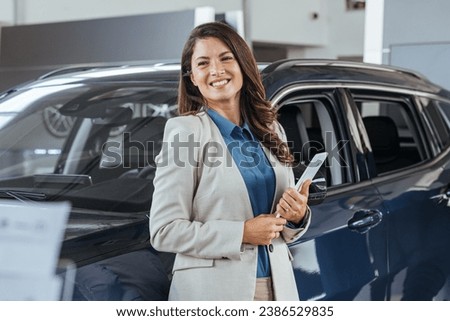 Smiling saleswoman holding tablet PC while looking at camera at new car showroom. Professional car dealer posing proudly at auto showroom, smiling to the camera, holding tablet PC.