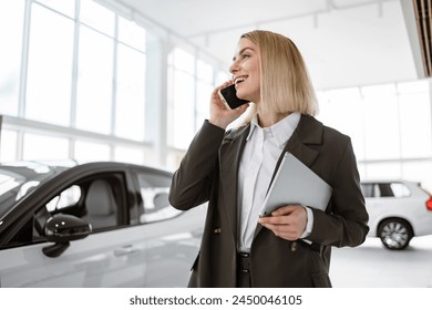 Smiling saleswoman smartphone while having business call at car showroom. Car dealer holding tablet pc in automobile showroom. Professional confident sales person working in modern car dealership. - Powered by Shutterstock