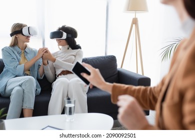 Smiling Same Sex Couple In Vr Headsets Holding Hands On Sofa Near Blurred Psychologist With Smartphone