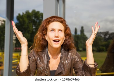 Smiling Sarcastic Woman Sitting In Outdoor Coffee Shop, Rolling Eyes, Gesturing. Red Curly Haired Young Woman In Casual Posing In Cafe. Sarcasm Or Relief Concept