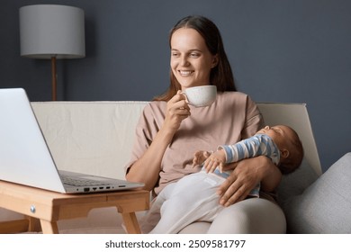 Smiling satisfied mother with her baby using on her laptop while drinking coffee and watching movie sitting on sofa in living room enjoying mug of tea while working on her computer - Powered by Shutterstock