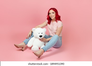 smiling, satisfied woman in jeans with red hair with a large white teddy bear sitting on the floor , isolated on a pink background.